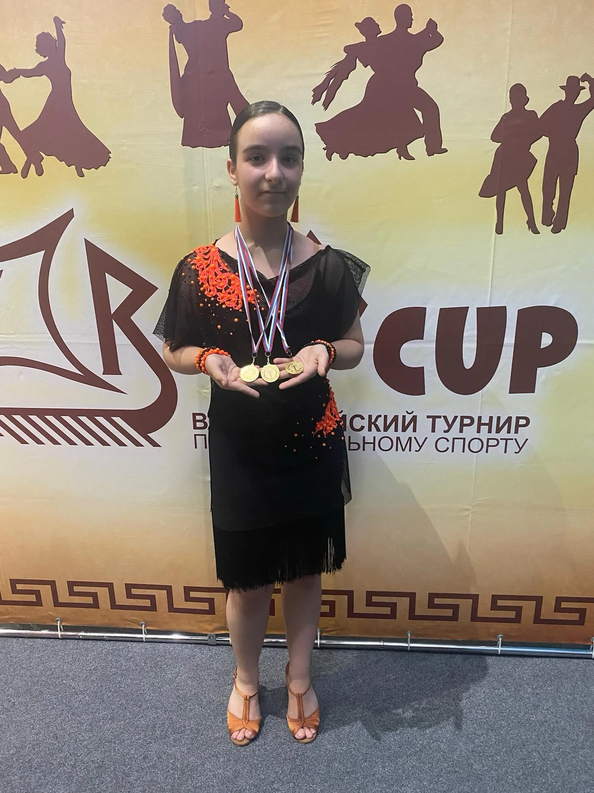 Ева в чёрно-оранжевом танцевальном платье с медалями у баннера турнира