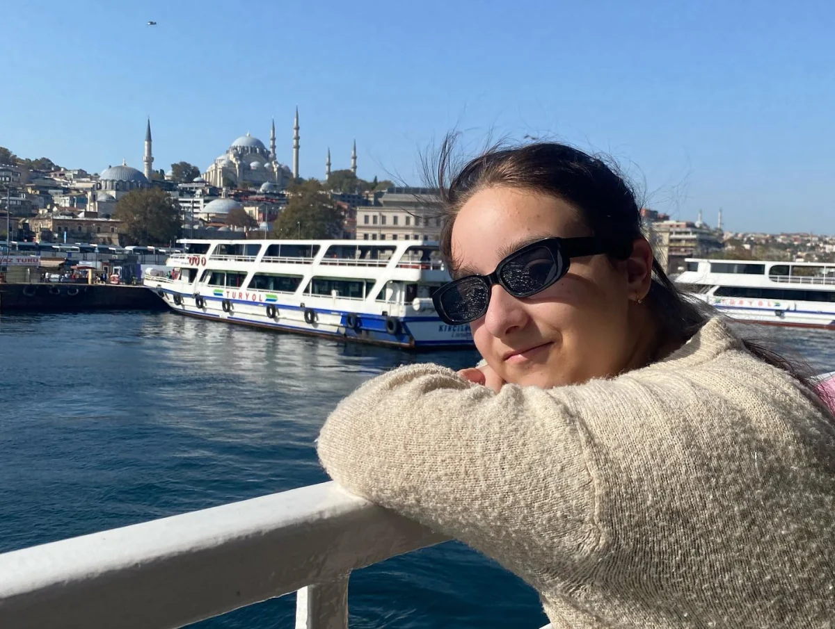 Улыбающаяся Ева в очках опирается на борт парома, на фоне вода и набережная Стамбула